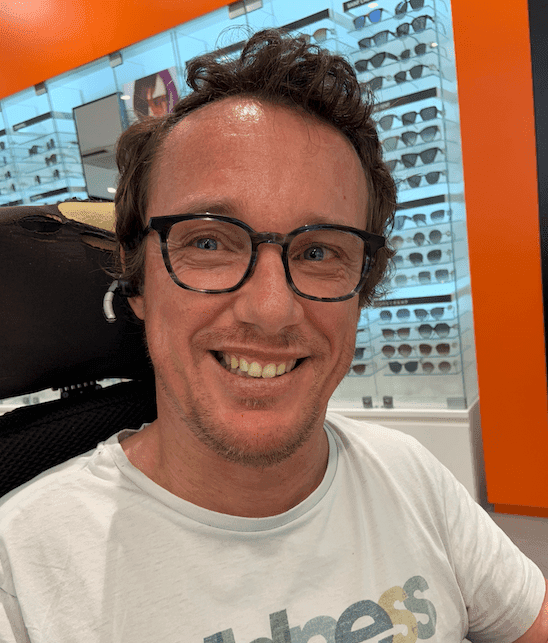 Portrait de Jonathan Dupire souriant, un homme caucasien portant des lunettes à monture noire. Il a des cheveux courts et bouclés et porte un t-shirt blanc. Jonathan est assis dans son fauteuil électrique avec un arrière-plan des présentoirs de lunettes.